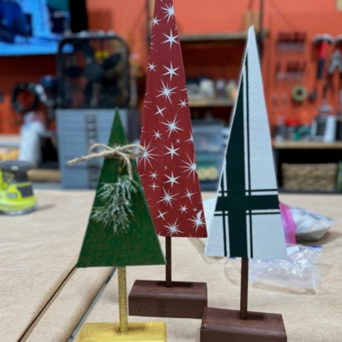 Three wooden Christmas tree figurines on stands with a blurred workshop background