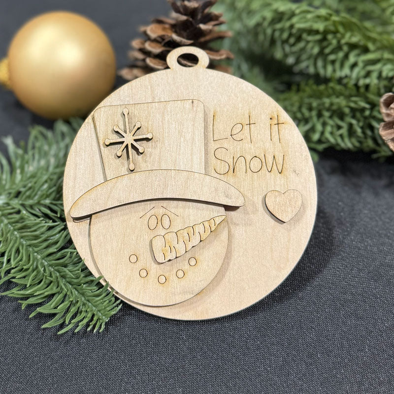 Wooden ornament with snowman design and 'Let it Snow' text, surrounded by Christmas decorations.