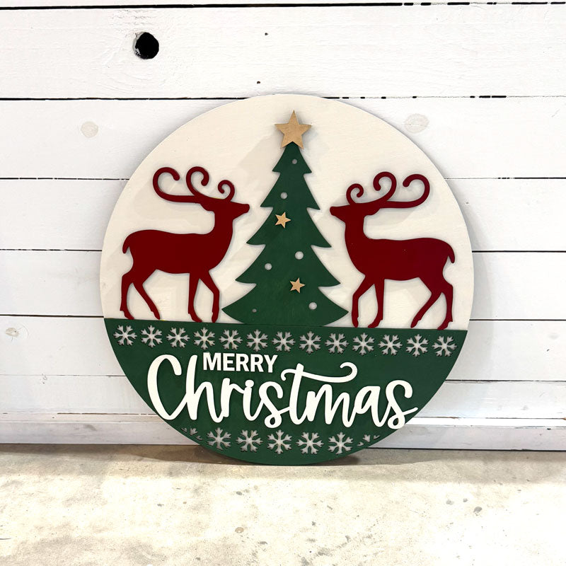 Round Christmas ornament with reindeer, tree, and 'Merry Christmas' text on a white wooden background.
