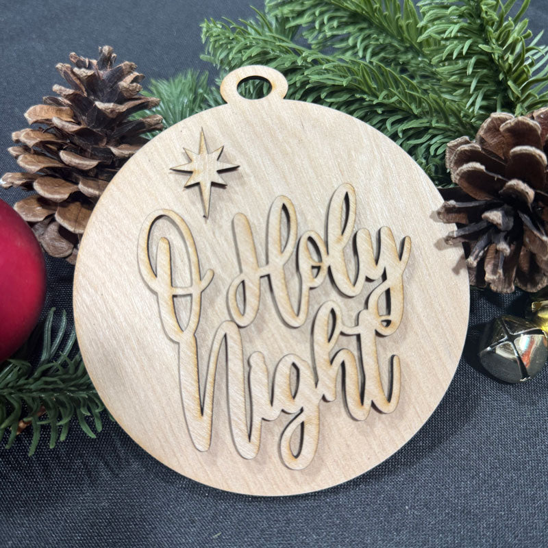 Wooden ornament with 'O Holy Night' text surrounded by pine cones and a red ball on a dark background
