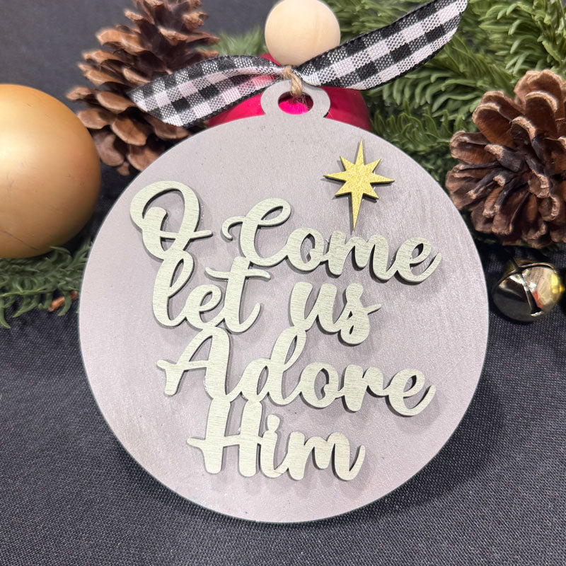Decorative Christmas ornament with 'O Come Let Us Adore Him' text and star design, surrounded by pine cones and a gold ball.