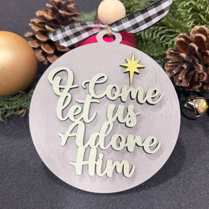 Decorative Christmas ornament with 'O Come Let Us Adore Him' text and star design, surrounded by pine cones and a gold ball.