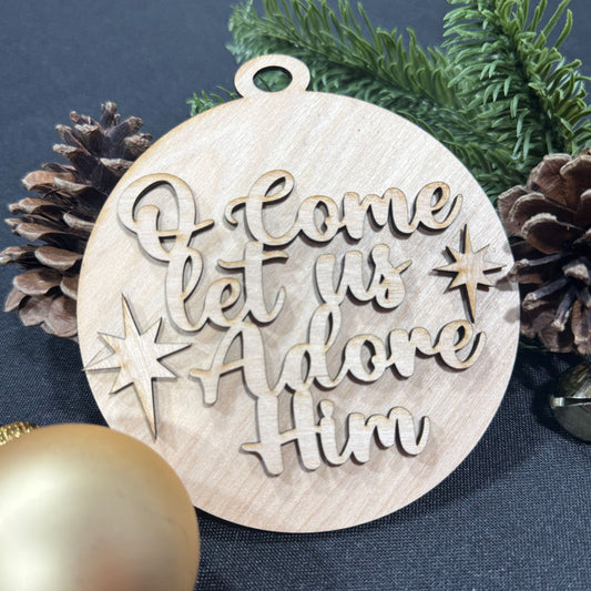 Wooden Christmas ornament with 'Come let us Adore Him' text surrounded by pine cones and a gold ball on a dark blue background.