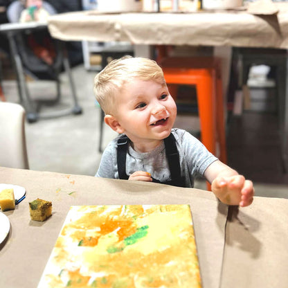 A young child sitting at a table engaging with a craft project, with paint and art supplies around.