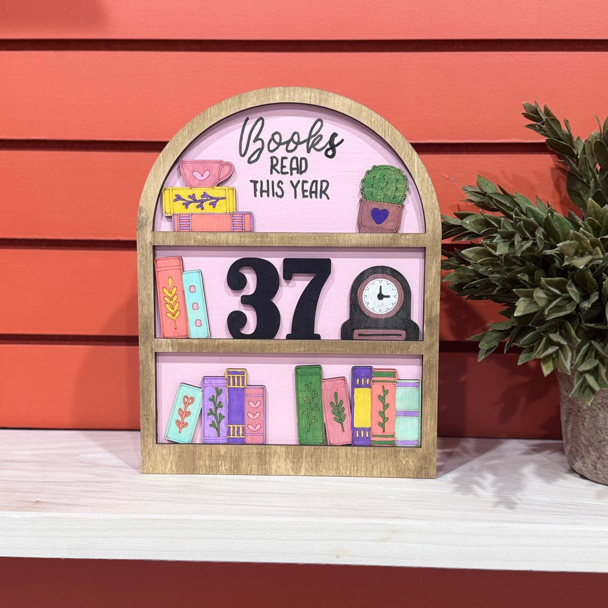 A decorative wooden shelf sign with a pink face, painted book illustrations, the words 'Books Read This Year,' and a large black number 37, displayed on a white shelf against a coral-red slatted wall with a potted plant beside it.
