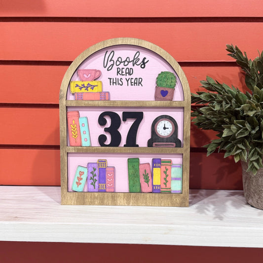 A decorative wooden shelf sign with a pink face, painted book illustrations, the words 'Books Read This Year,' and a large black number 37, displayed on a white shelf against a coral-red slatted wall with a potted plant beside it.