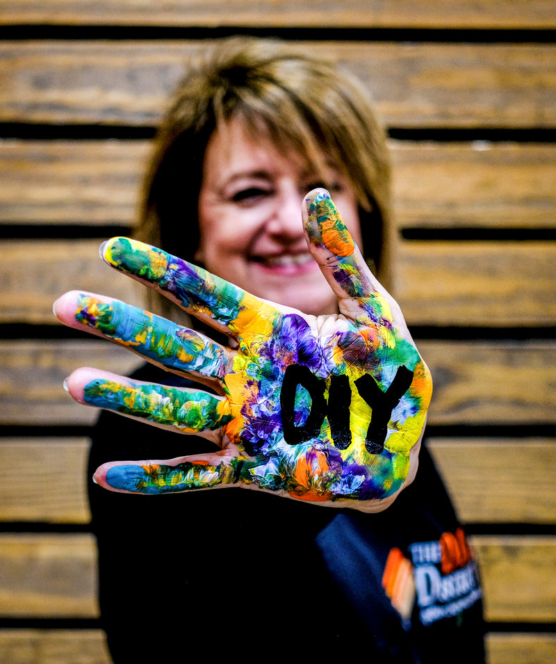 A smiling woman holds up her hand, which is covered in colorful paint splatters and has the word 'DIY' written in black marker, against a wooden slat wall background.