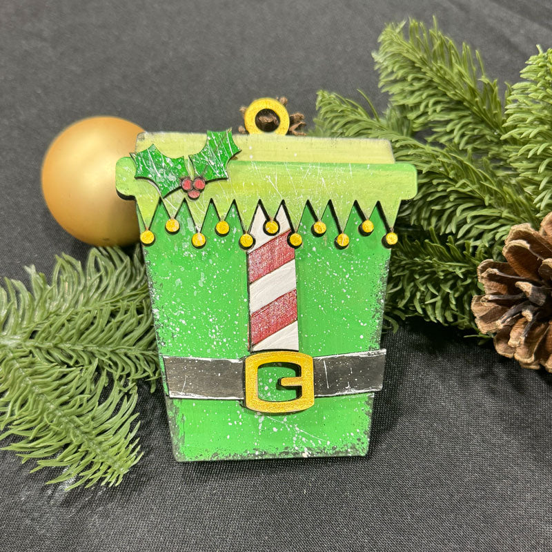 Decorative Christmas ornament coffee cup with an elf design, surrounded by greenery and pine cones.