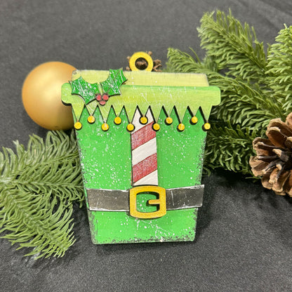 Decorative Christmas ornament coffee cup with an elf design, surrounded by greenery and pine cones.