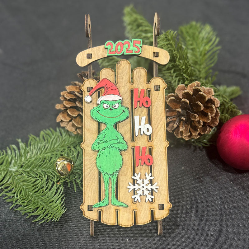 Christmas ornament with Grinch design, 'Ho Ho Ho' text, and '2025' on a wooden background.