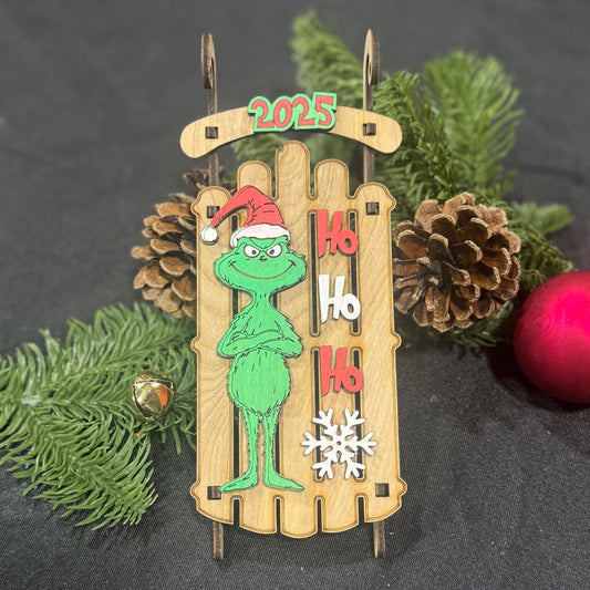 Christmas ornament with Grinch design, 'Ho Ho Ho' text, and '2025' on a wooden background.