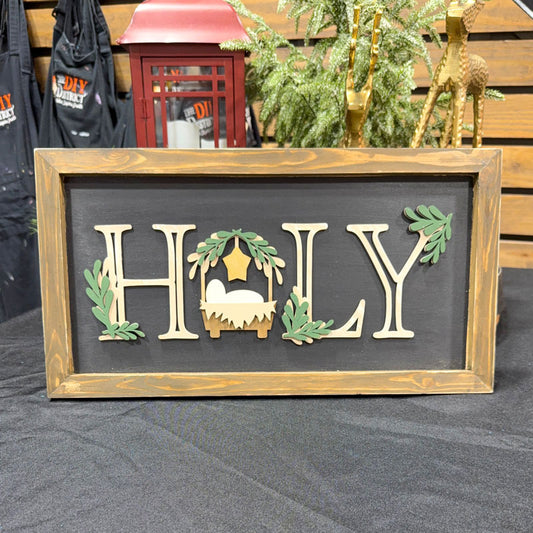 Decorative sign with 'HOLY' letters, featuring a nativity scene, on a wooden stand.