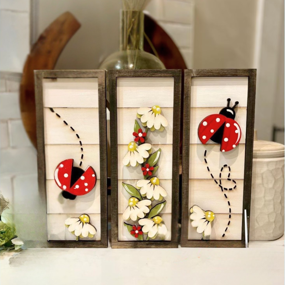 Three wooden panels with ladybug and flower designs on a white surface.