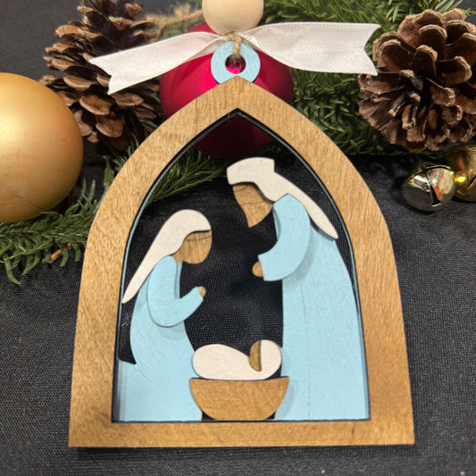 Decorative nativity scene ornament with wooden frame and Christmas decorations in the background.