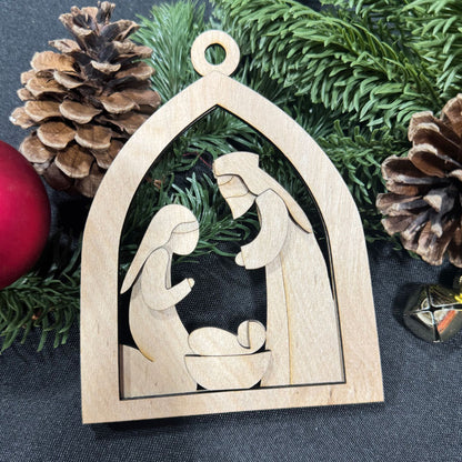 Wooden nativity scene ornament with pine cones and greenery on a dark background
