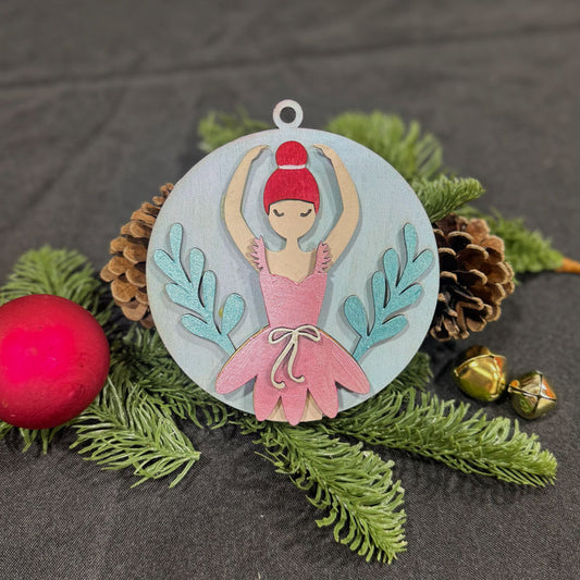 Decorative ornament with a ballerina design on a dark surface with greenery and pine cones.