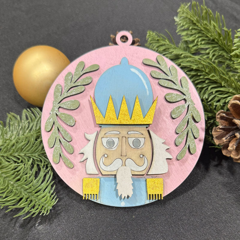 Decorative ornament with a nutcracker design on a gray background with greenery and a gold ball.