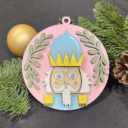 Decorative ornament with a nutcracker design on a gray background with greenery and a gold ball.