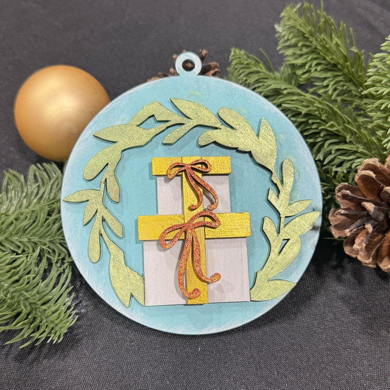 Decorative Christmas ornament with a gift and wreath design on a dark surface with greenery and a gold ornament.