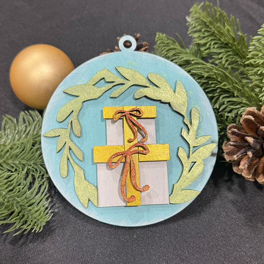 Decorative Christmas ornament with a gift and wreath design on a dark surface with greenery and a gold ornament.