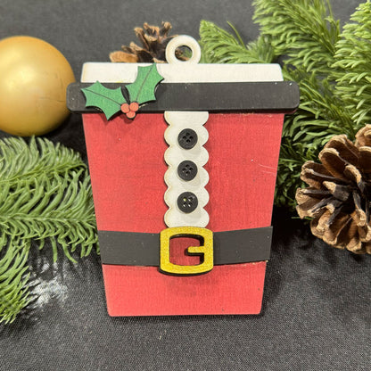 Red cup-shaped Christmas ornament with buttons and holly leaves on a dark background with greenery.