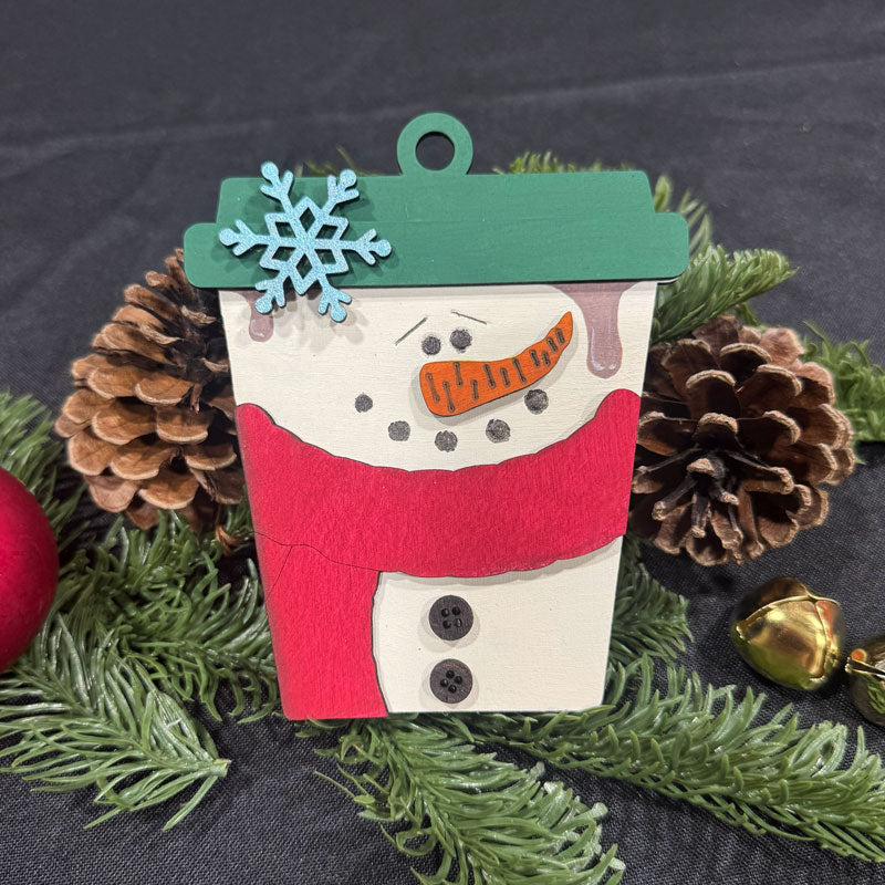 Wooden snowman ornament with a green hat and red scarf, surrounded by pine cones and greenery on a dark background.