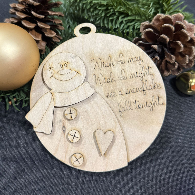 Wooden ornament with snowman design and text, surrounded by Christmas decorations on a dark surface.