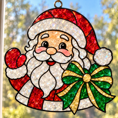 A hanging Christmas suncatcher featuring a glittery Santa Claus face and beard, red hat, red mitt, and a green and gold bow, photographed against a blurred outdoor window background.