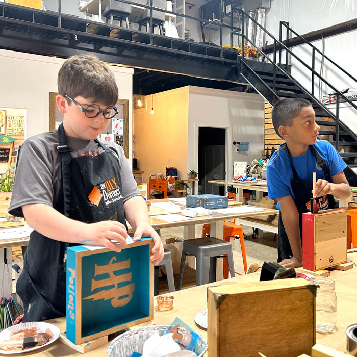 Two children participating in a DIY crafting activity, creating items on workbenches in a workshop setting.