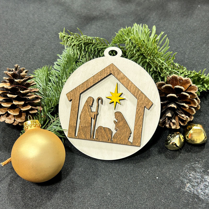Decorative Christmas ornament with nativity scene design, surrounded by pine cones and a gold ball on a dark background.