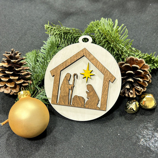 Decorative Christmas ornament with nativity scene design, surrounded by pine cones and a gold ball on a dark background.